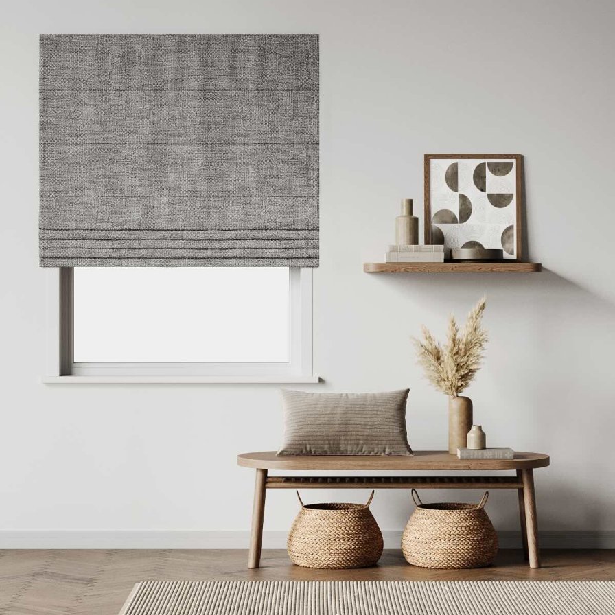 A grey textured roman blind covering a window in a styled living room with a floating shelf and woven baskets.