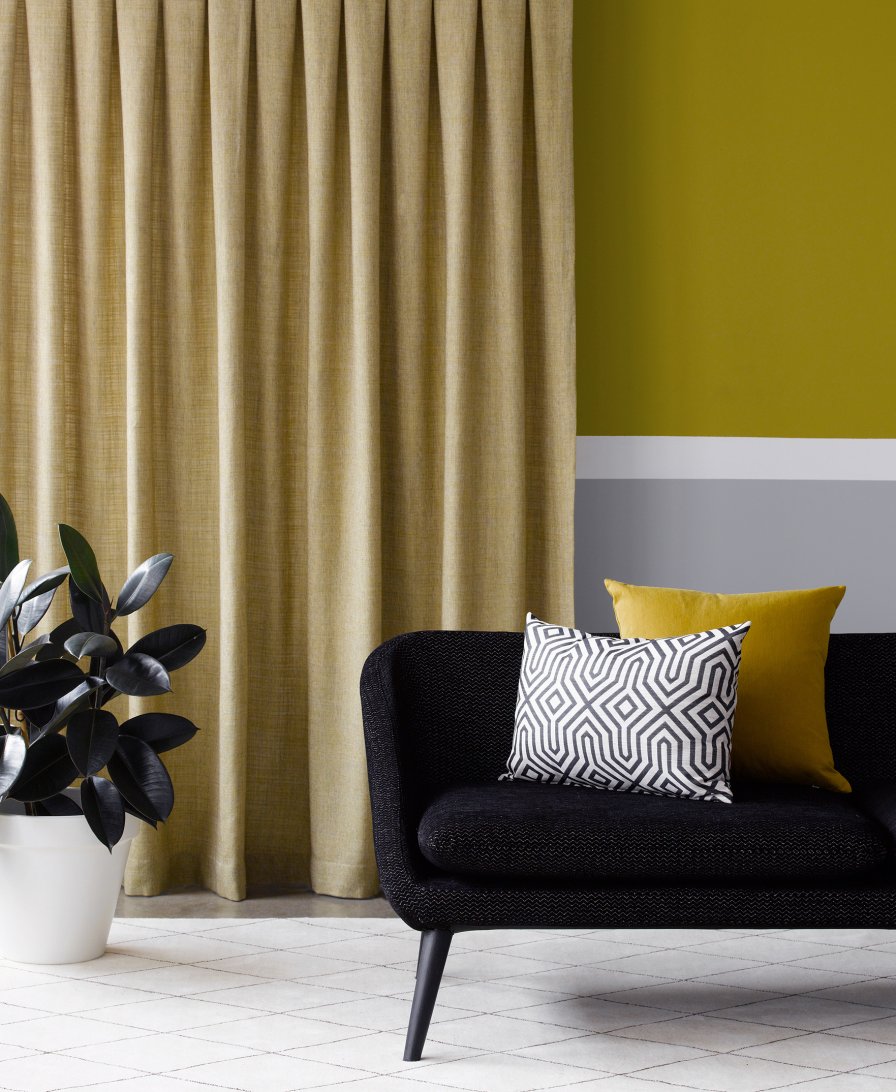 Pale sage pleated curtains beside a black sofa with patterned cushions against an olive-green wall.
