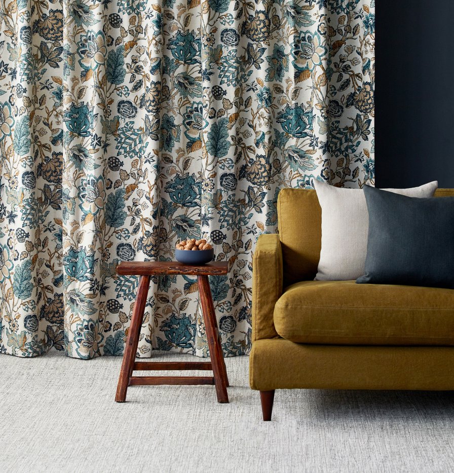 Curtains with a blue-green botanical print beside a mustard-yellow armchair and wooden side table.