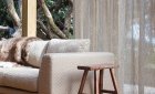 Natural taupe sheer curtains at a window beside a sofa and wooden stool, with trees outside.