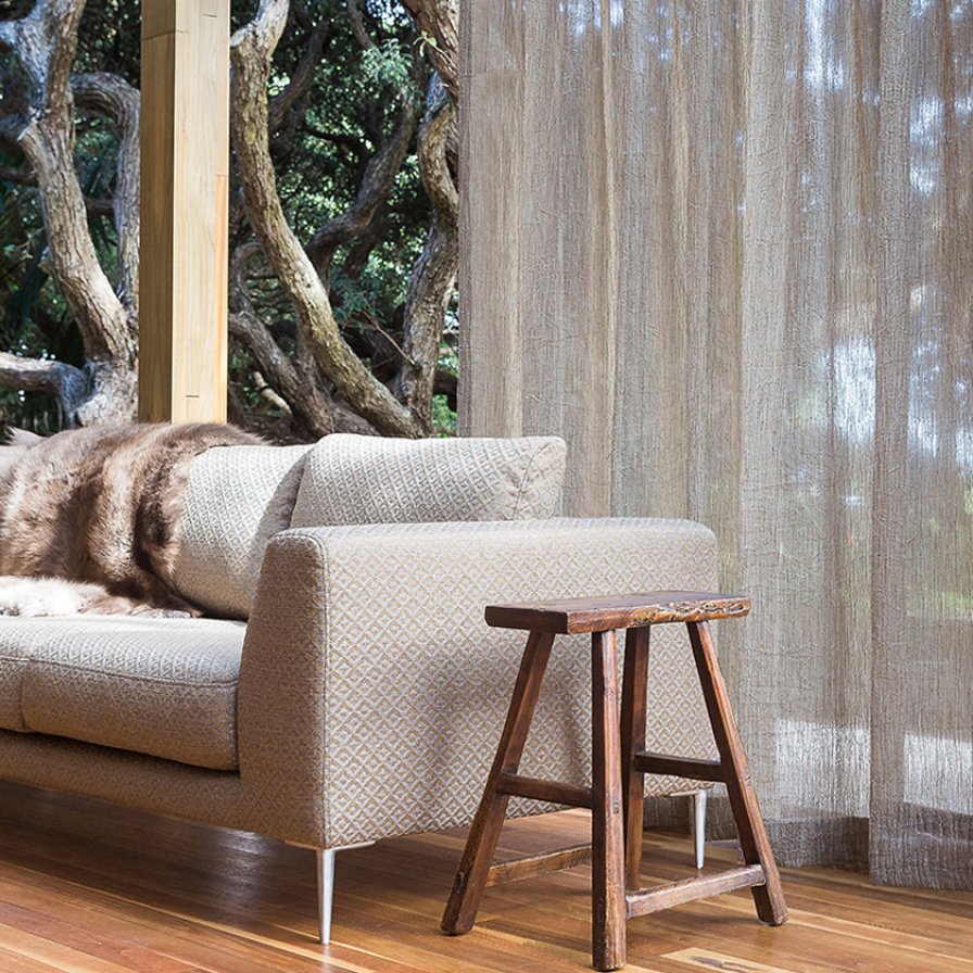 Natural taupe sheer curtains at a window beside a sofa and wooden stool, with trees outside.