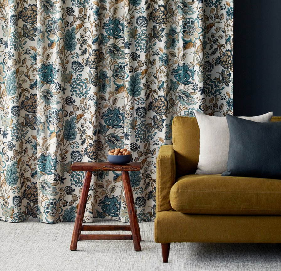 Cream curtains with a teal, grey and black botanical print beside a mustard-gold sofa.
