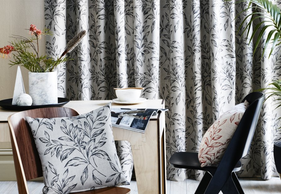 White and dark grey leaf-patterned curtains and cushion beside a wooden dining table.