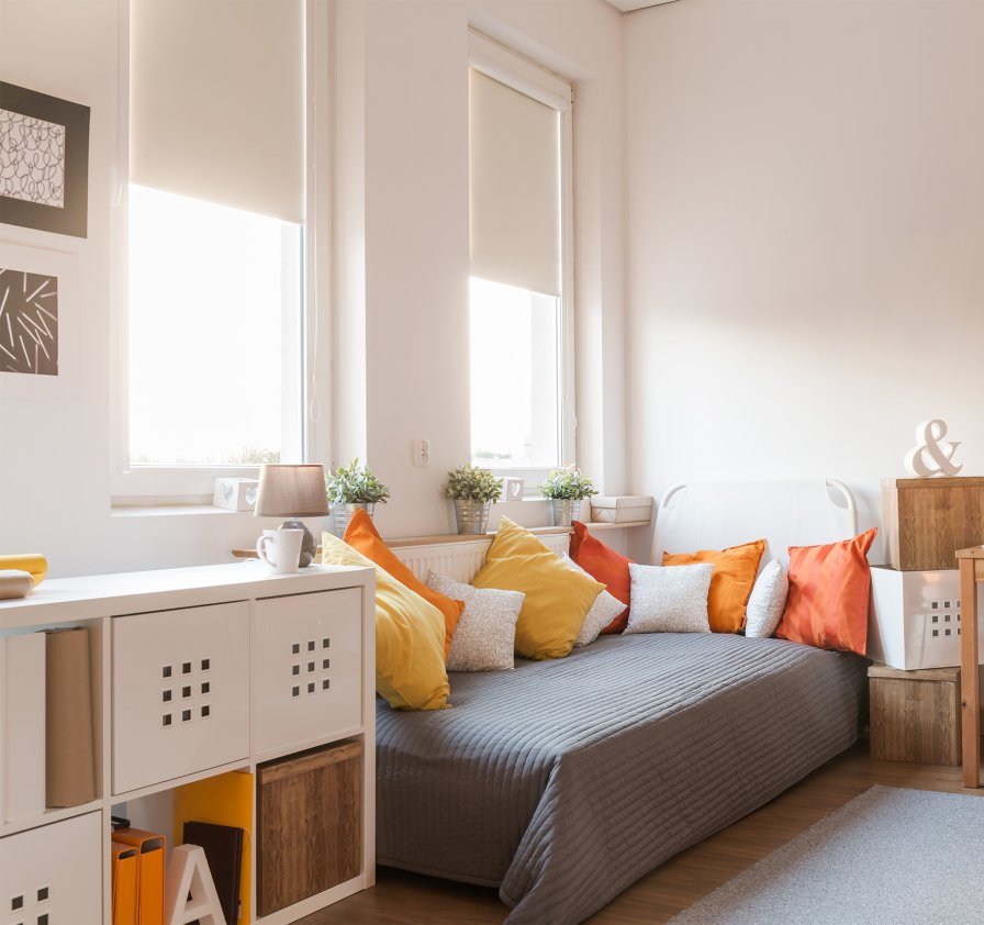 Bedroom with cream thermal roller blinds at two tall windows beside a bed with warm cushions.