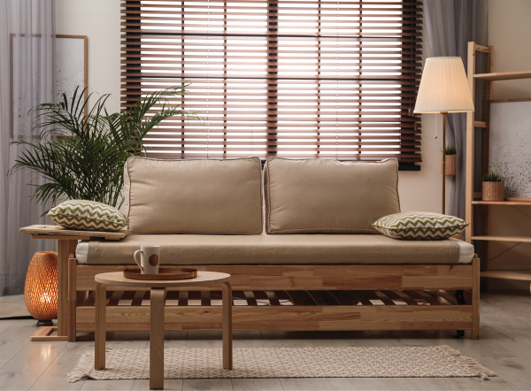 Mid-brown wood venetian blinds on a window behind a wooden sofa with cushions and a plant.