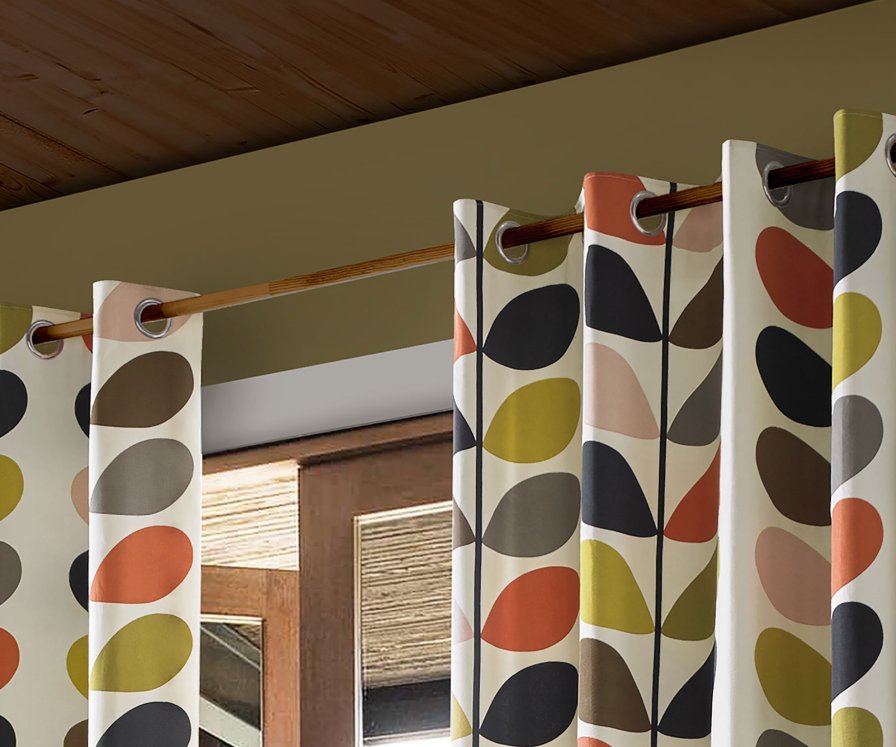 Cream eyelet curtains with a rust, navy and olive geometric pattern on a wooden pole.