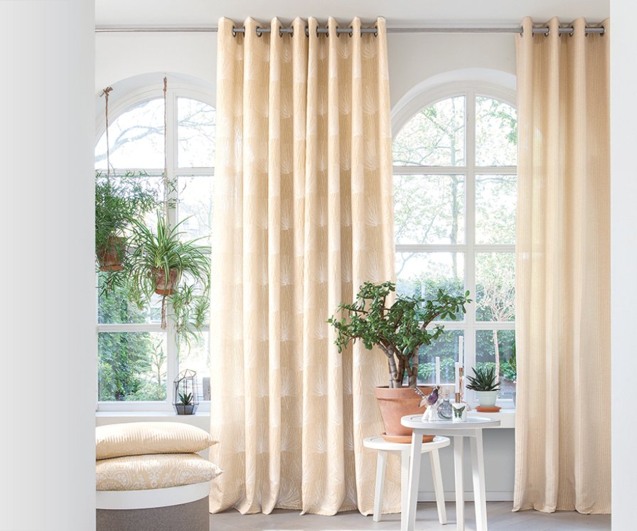 Cream eyelet curtains on poles flanking arched windows in a bright, plant-filled living room.