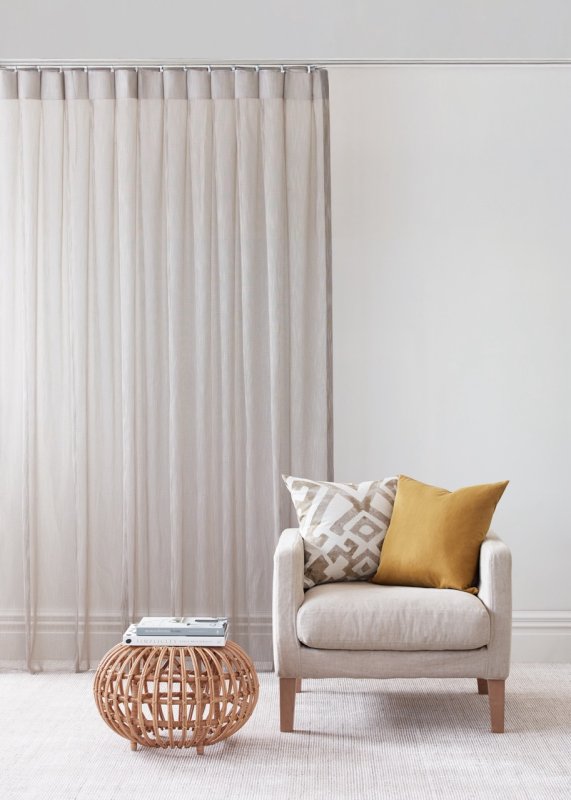 Cream sheer pencil pleat curtains with a taupe stripe on a chrome pole beside a cream armchair.