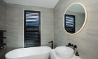 Black plantation shutters on a tall window in a grey-tiled bathroom with a freestanding tub.