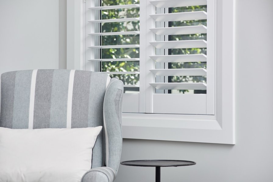 White plantation shutters on a window beside a grey striped armchair.