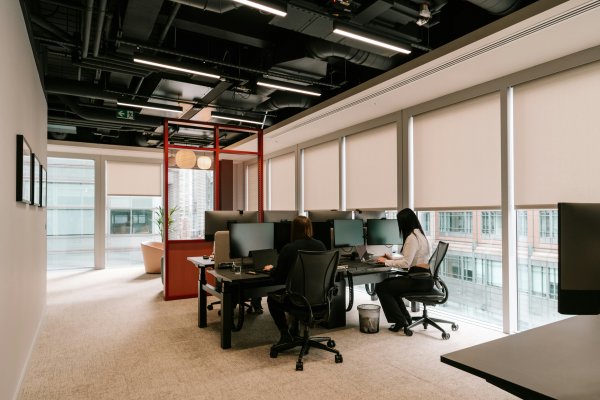 Cream roller blinds along the windows of an open-plan office with people working at desks.