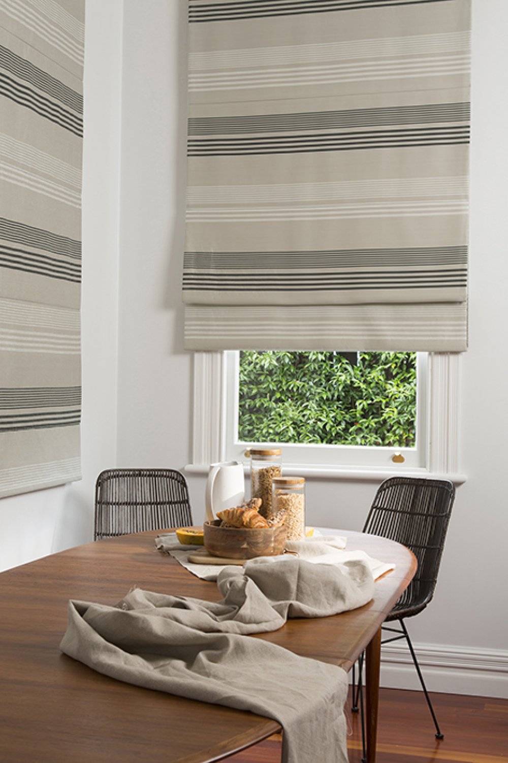 Cream and charcoal striped roman blinds in a dining room above a window with rattan chairs.