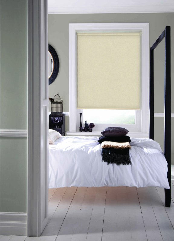 Pale yellow roller blind in a minimalist bedroom with white bedding and dark accents.