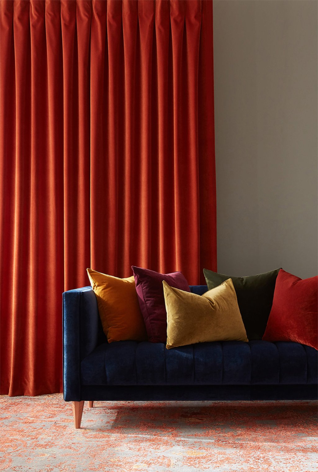 Burnt orange pencil pleat curtains beside a dark blue velvet sofa with colourful cushions.