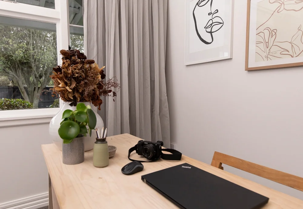 A workspace by a window with grey pleated curtains, a desk, plants and artwork.