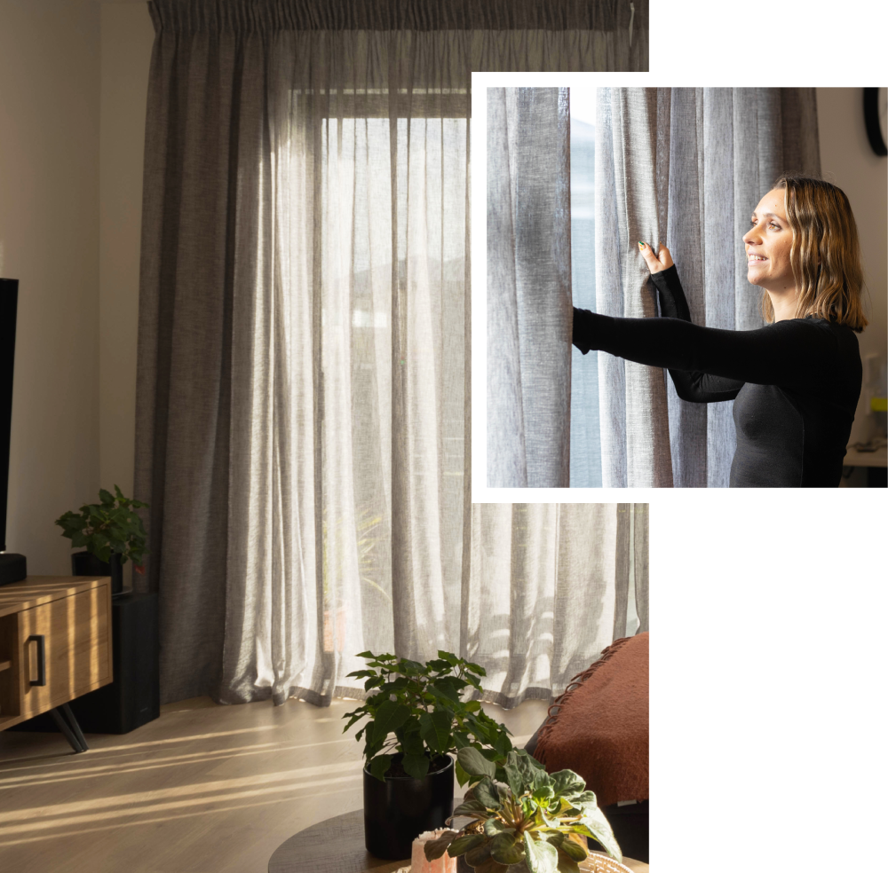 A woman touching a grey textured curtain in a neutral-toned living room.