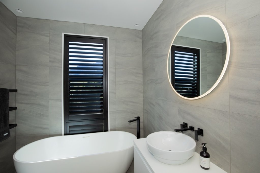 Black plantation shutters on a tall window in a grey-tiled bathroom with a freestanding tub.