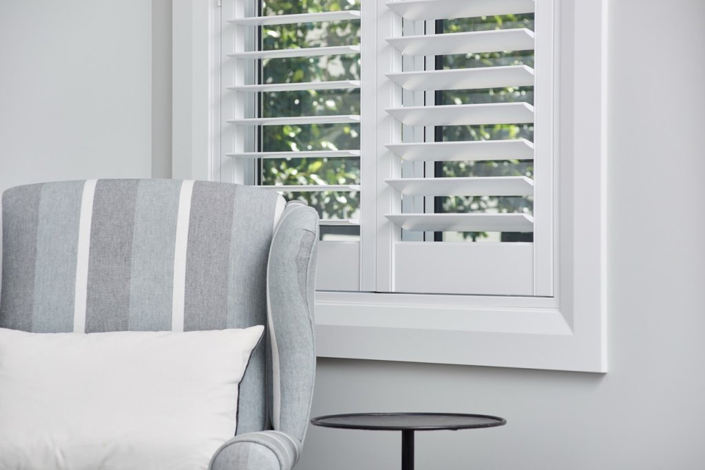 White plantation shutters on a window beside a grey striped armchair.