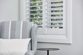 White plantation shutters on a window beside a grey striped armchair.