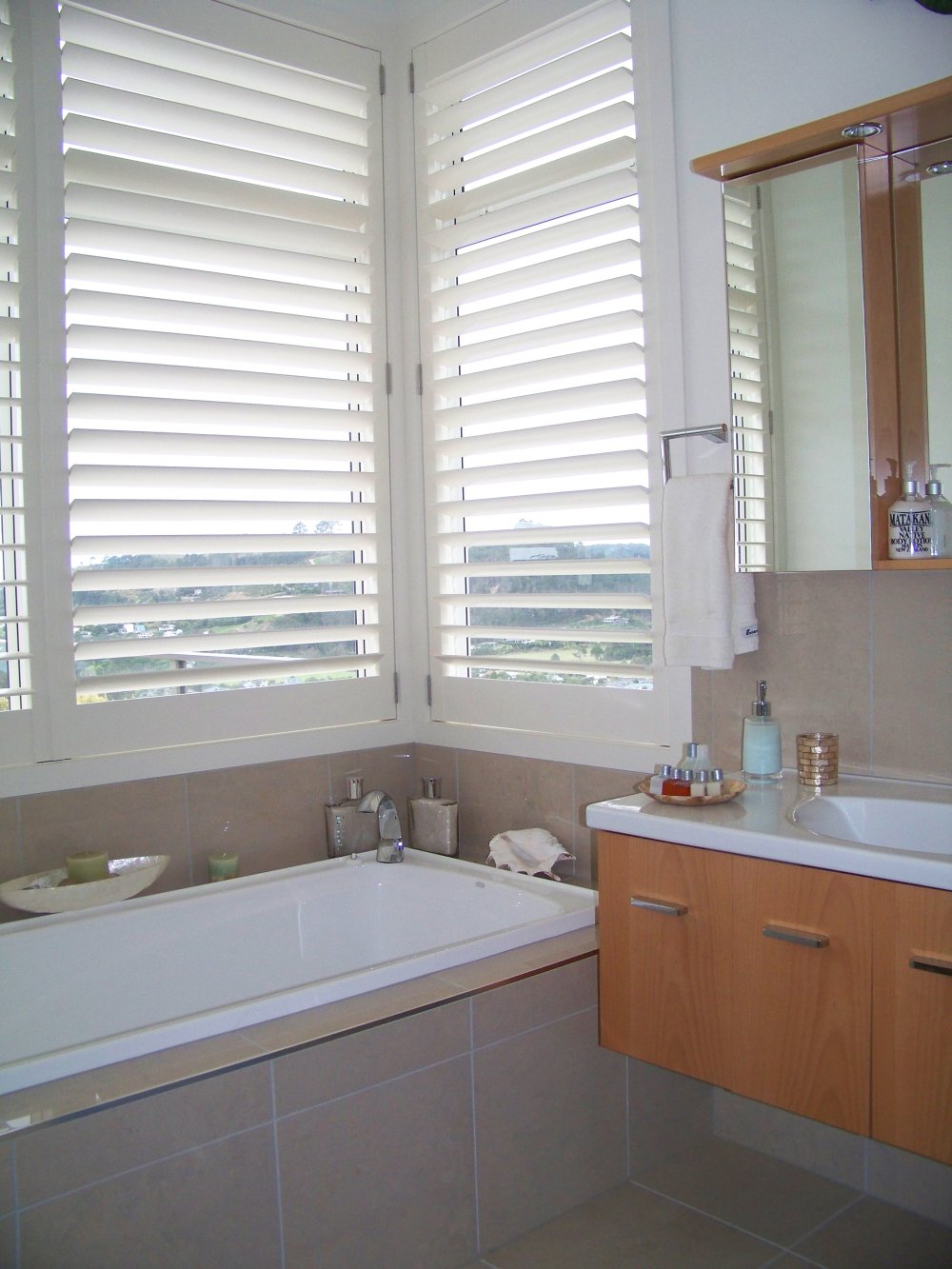 White shutters above a bath in a bathroom with tan tiled walls and a wooden vanity.