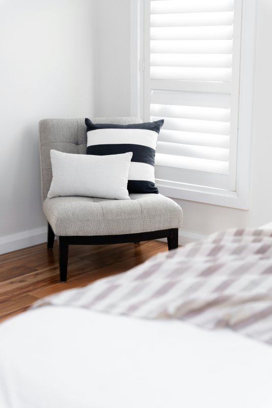 White horizontal shutters on a bedroom window beside a grey chair and striped bedding.