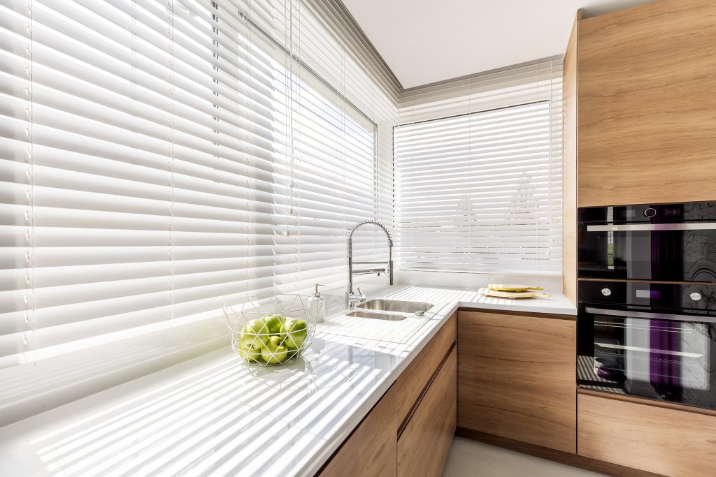 Modern kitchen with white horizontal blinds and wooden cabinetry beside a sink.